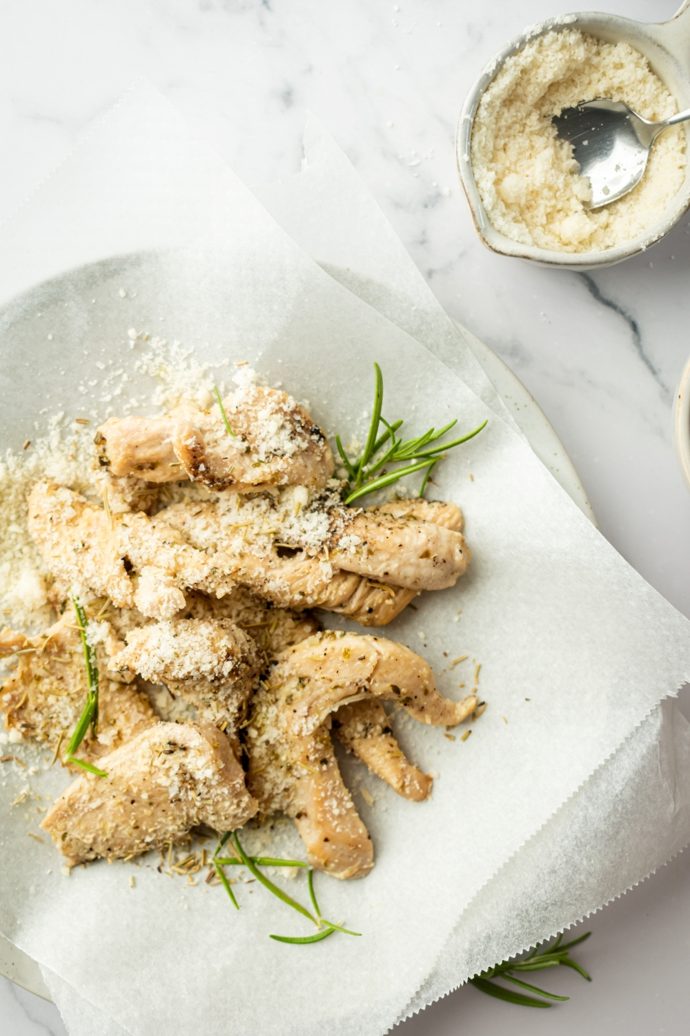 Chicken tenders with Parmesan cheese on top on a piece of parchment paper. Behind them is a small bowl with Parmesan cheese and a spoon in it on a white counter.