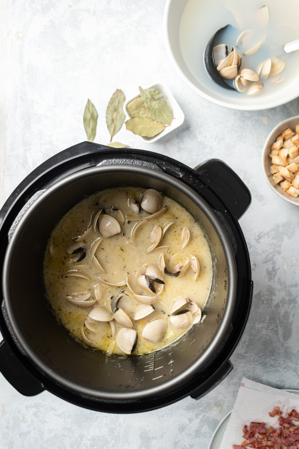 An instant pot filled with clams and other clam chowder ingredients. Behind it on the gray counter is part of a white bowl filled with clams in water.