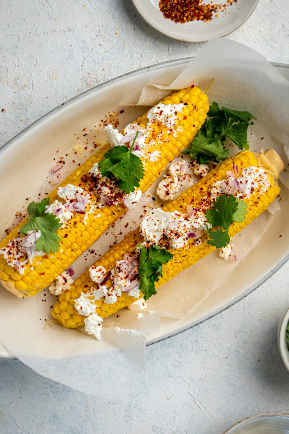 A white bowl wine with parchment paper with two pieces of corn on the cob on top. There is a Mexican queso fresco and cilantro on top of the corn.
