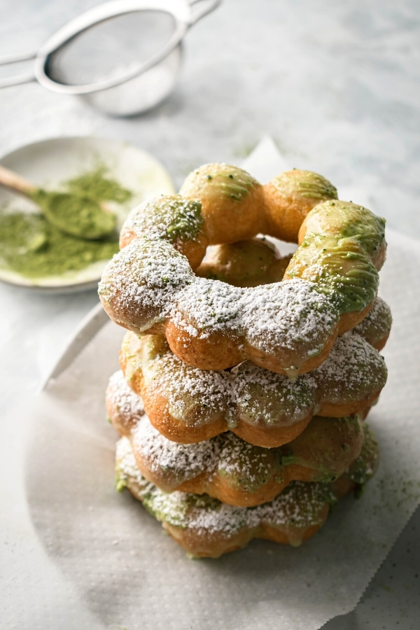 Four mochi donuts stacked on top of each other on a paper towel lined plate. Behind the stack is a small white plate with matcha powder on it and a mesh sieve with powdered sugar in it behind that.