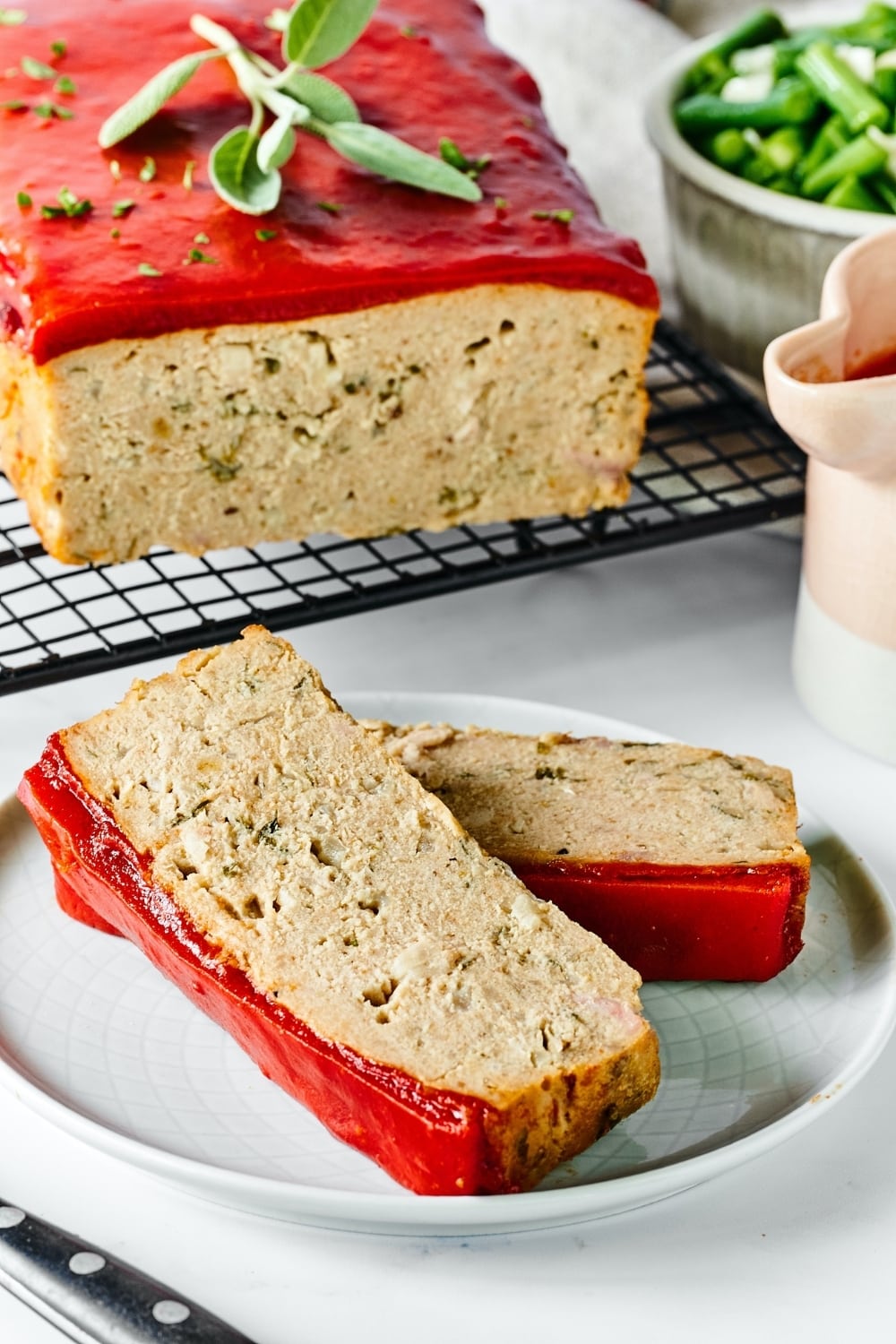 A white plate with two slices of chicken meatloaf on it. Behind the plate is a black wire rack with part of the chicken meatloaf on it.