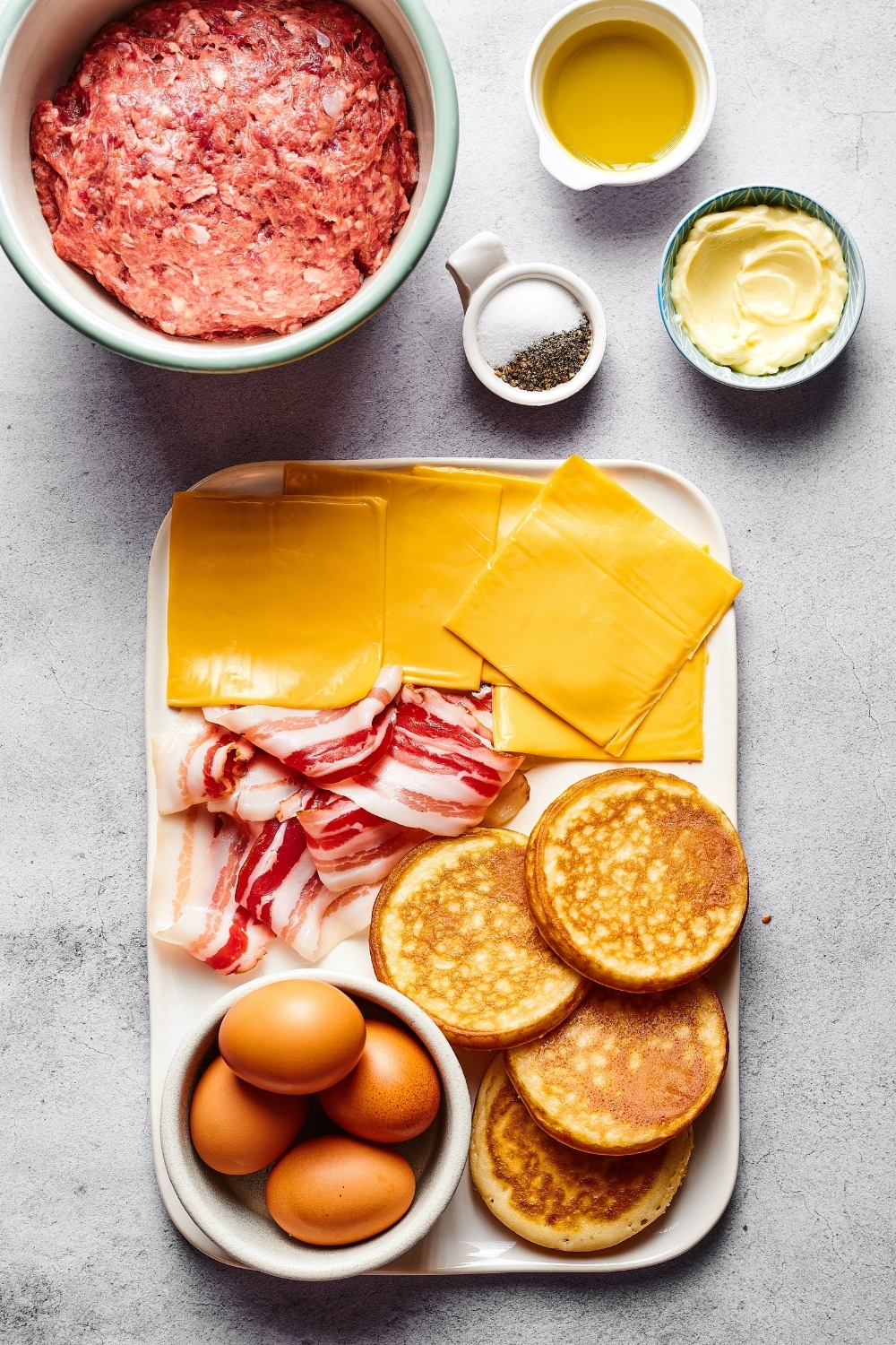 A rectangular white plate with four mcgriddle pancakes, a small white bowl with four eggs in it, a few strips of bacon, and a few slices of American cheese all on the plate. Behind that is a large bowl with raw ground beef and next to that small bowl of salt and pepper, small bowl of butter, and a small bowl of olive oil.