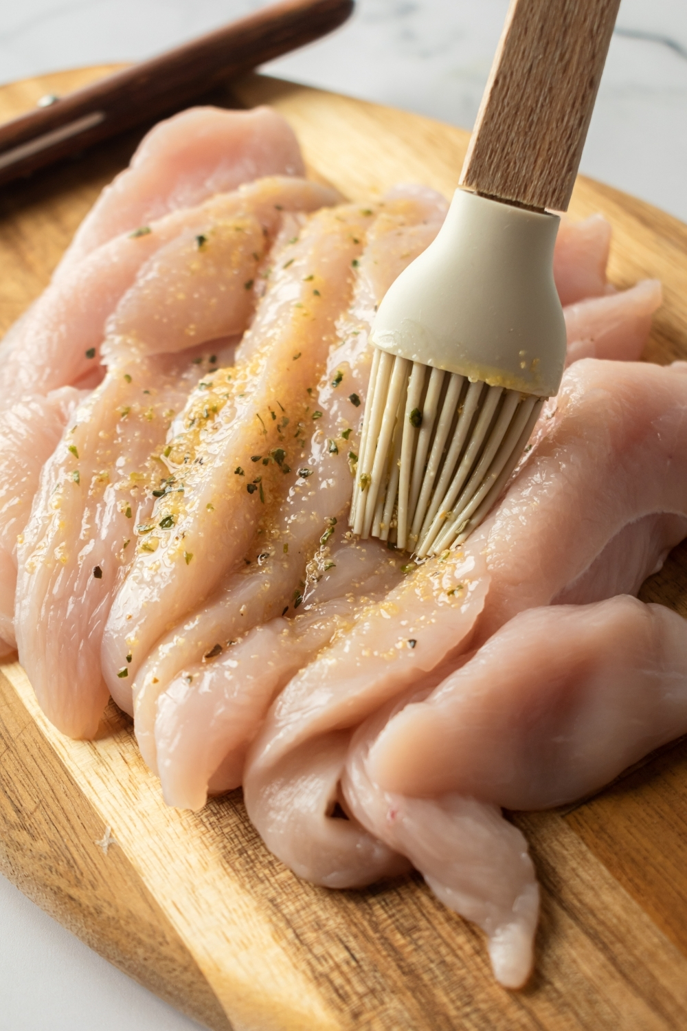 Raw chicken tenders on a wooden cutting board being brushed with an olive oil seasoning mixture.