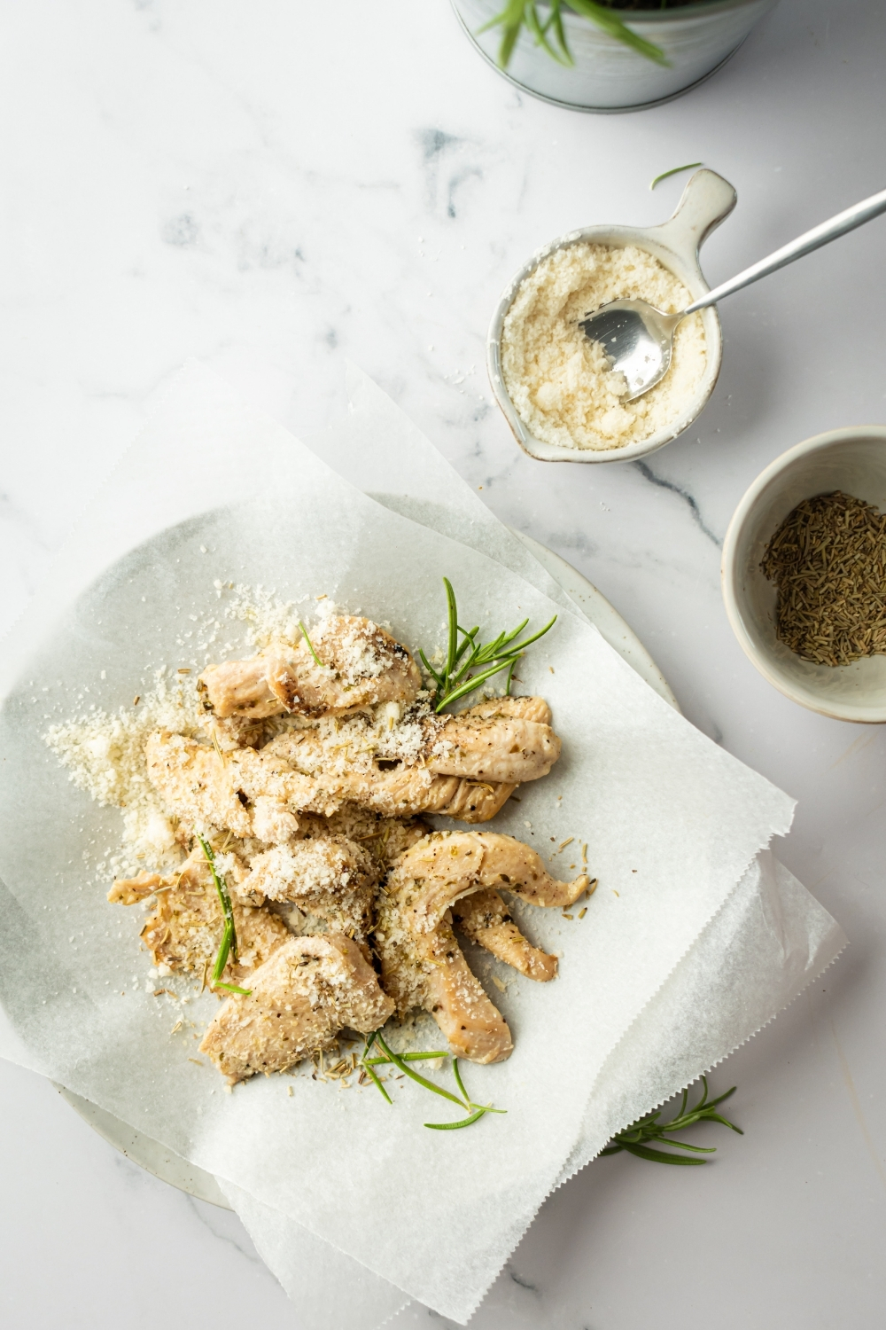 Chicken tenders with Parmesan cheese on top and a piece of parchment paper. Behind it is a small bowl of Parmesan cheese with a spoon in it on a white counter.