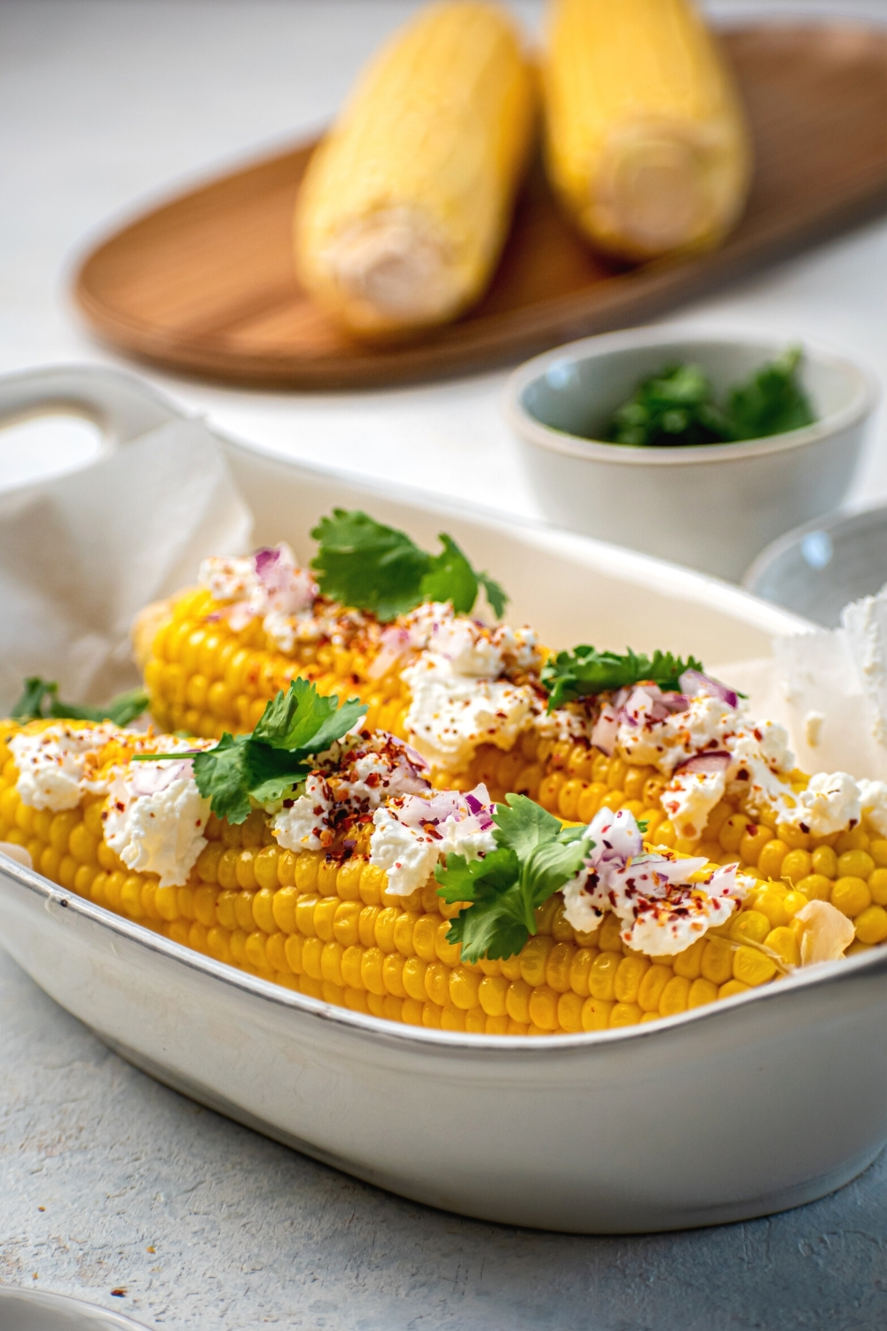 Two pieces of corn on the cob with Mexican queso fresco on top. Behind it is a bowl with cilantro and behind that a wooden board of two pieces of corn on the cob on it.