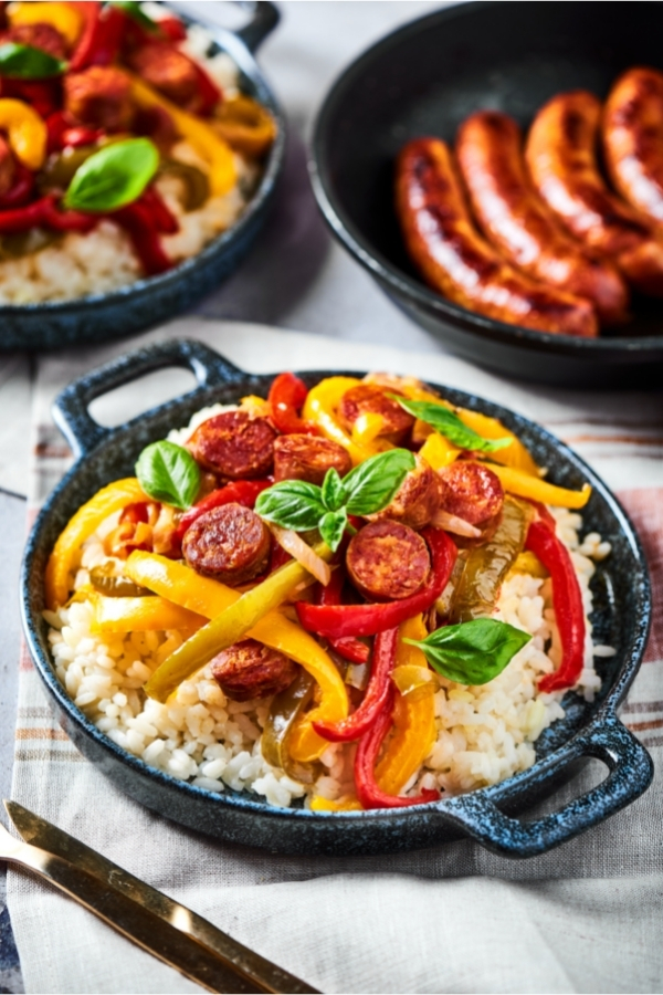 A bowl filled with white rice with peppers, onions, and slices of sausage on top. Behind that is part of a skillet with three cooked sausages on it and next to that is part of a bowl with rice, peppers, onions, and sausage.
