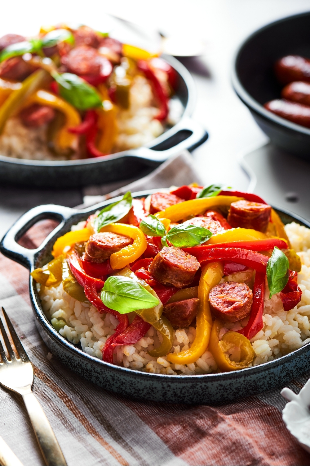 A bowl with sliced Italian sausage, red, green, yellow peppers and onions all on top of white rice. The bowl is on a plaid tablecloth and there is a fork next to the bowl. There is another bowl behind it with the same thing in it on a grey counter.