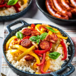 A bowl filled with white rice, sliced red, green, and yellow peppers and onions, and slices of sausage white rice. The bowl is on a plaid tablecloth and behind it is part of a pan with three cooked sausages on it next to that another bowl with rice, sausage, peppers and onions.