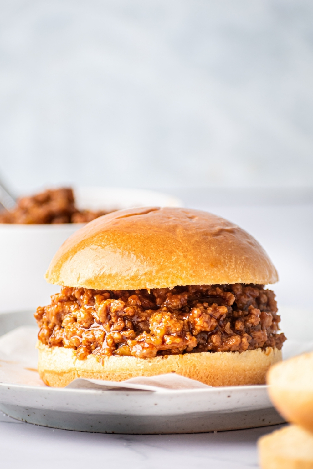 A sloppy Joe on a piece of parchment paper on a gray plate. Behind it is part of our bowl filled with sloppy Joes.