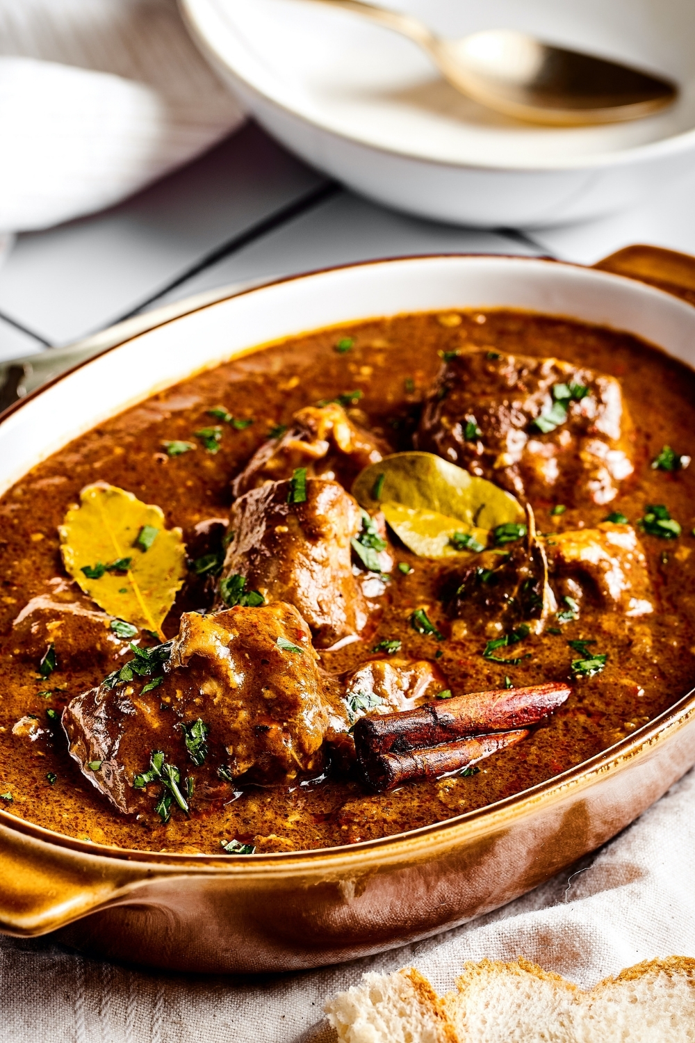 An oval serving dish that is gold on the outside and white on the inside filled with lamb vindaloo.