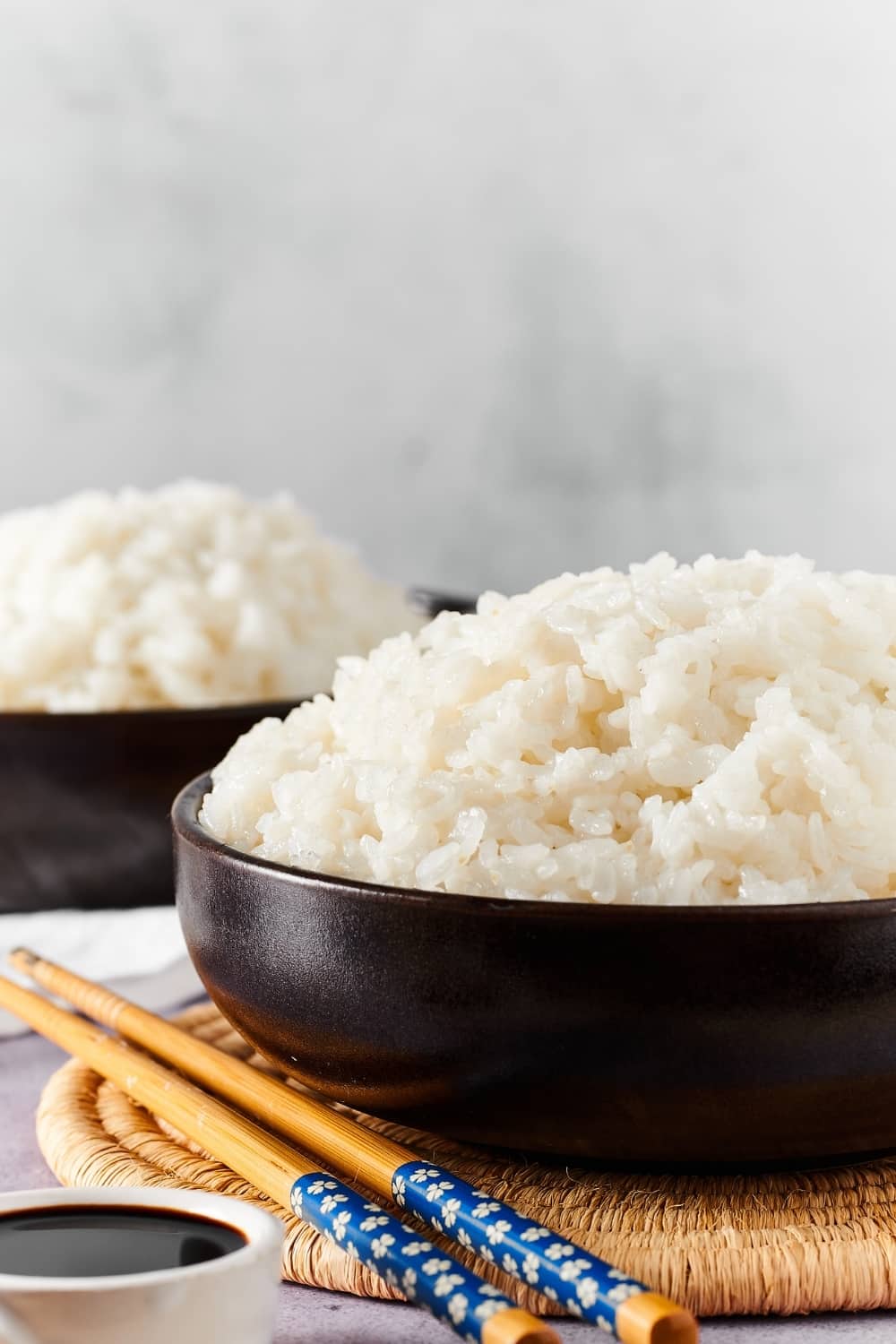 Part of a black bowl filled with sushi rice and a circular placemat. A pair of chopsticks to the left of the bowl on the placemat and in the background is a part of a black bowl filled with sushi rice.