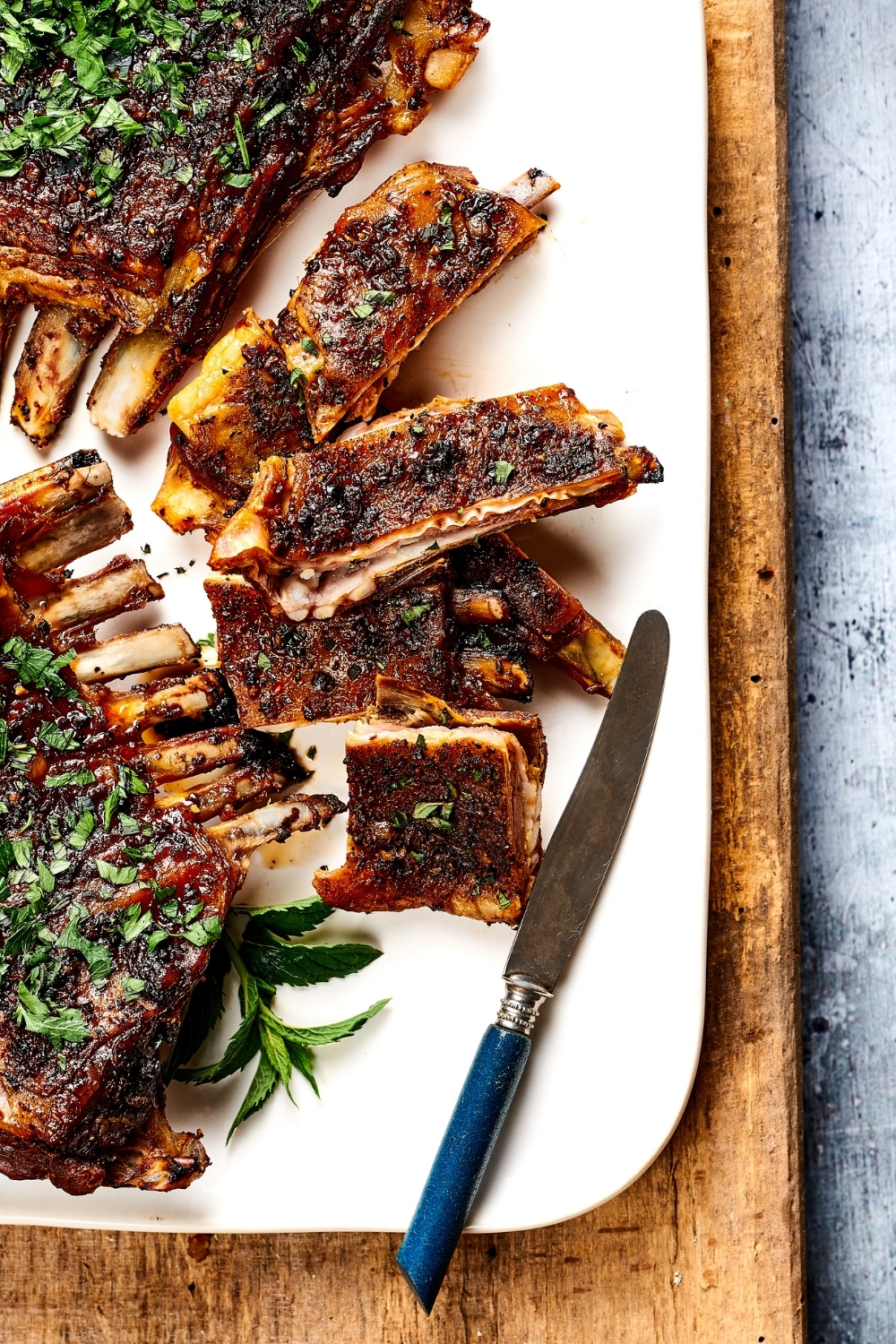 Part of a rectangular white plate with slices of lamb breast and a knife on it. To the left of the slices are parts of lamb breast with rib.