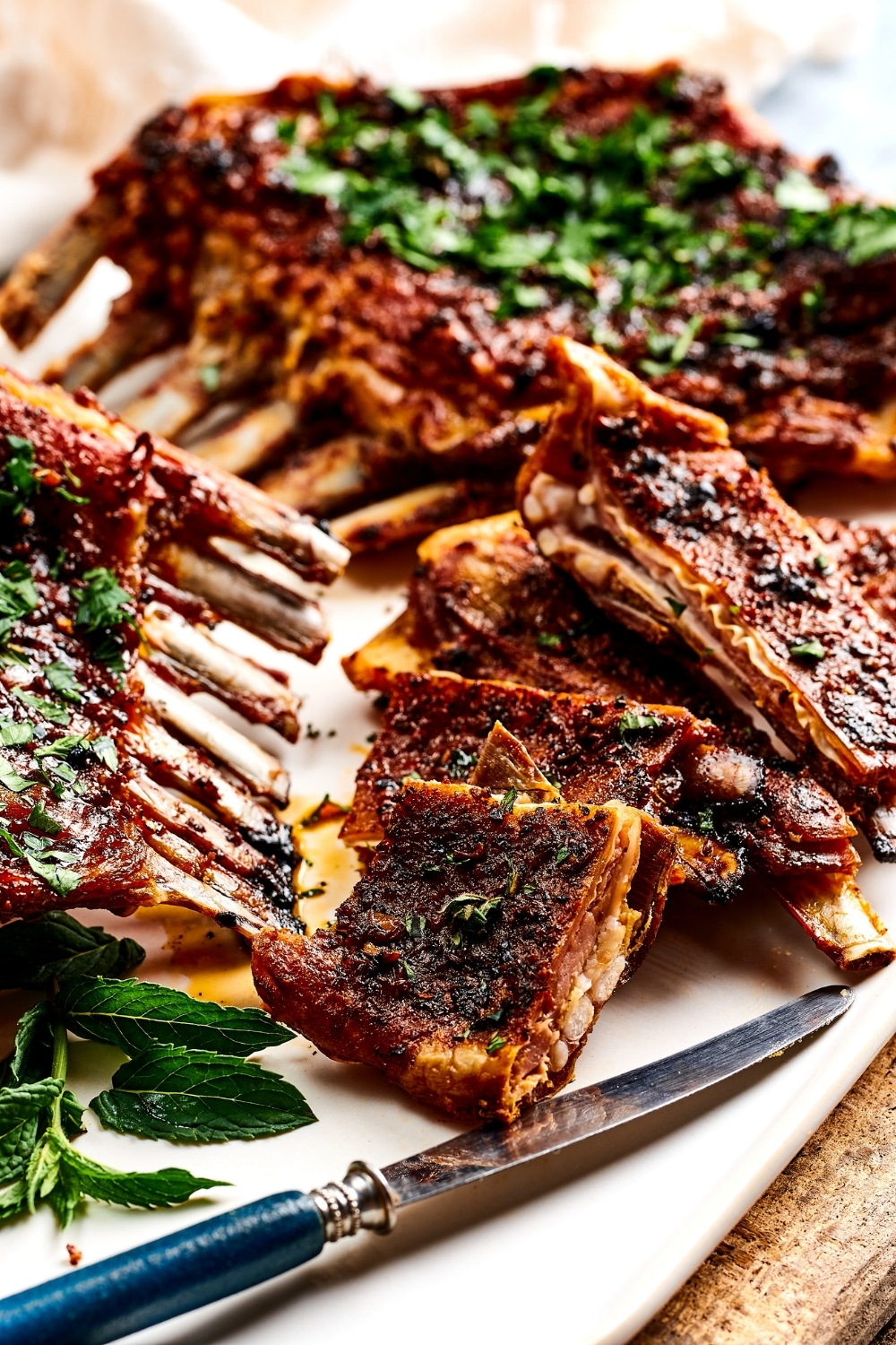 A few slices of lamb breast on top of one another on a white plate. Next to them is parts of the ribs of lamb breast and behind them as a whole lamb breast.