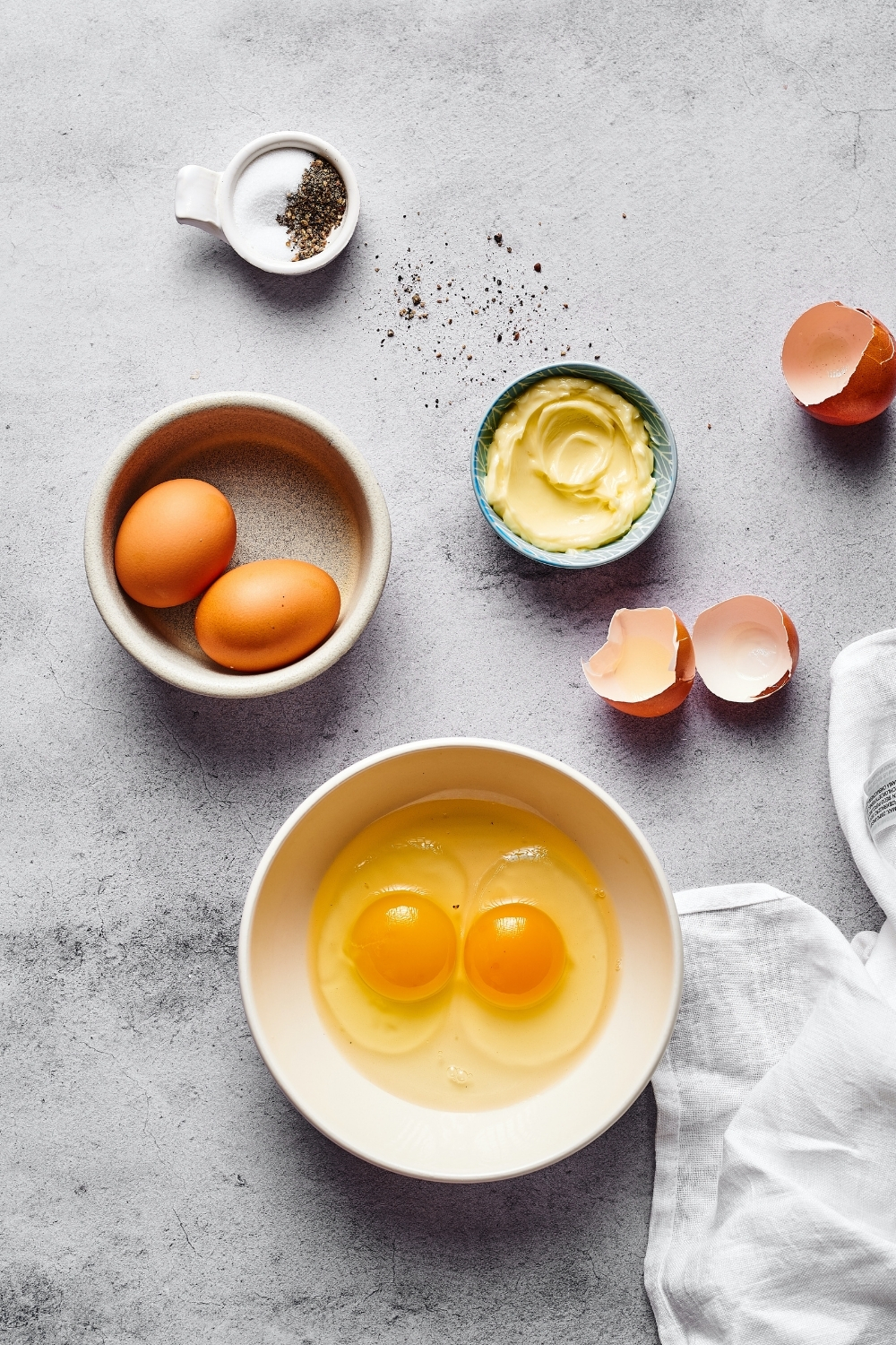 A whiteboard with two cracked eggs in it, a gray bowl in front of that with two whole eggs in it, to the right of that is a small bowl of butter, some egg shells, a small whiteboard salt and pepper.