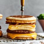 A sausage, meat, and cheese McGriddle on a wire rack on a white tablecloth on a wooden board on top of a counter. There is a knife through the top center of the McGriddle.