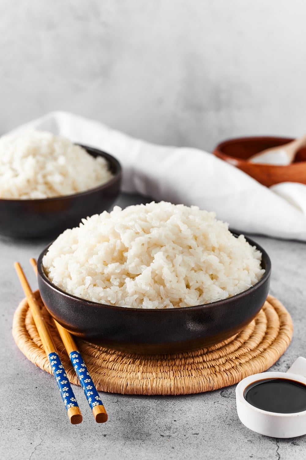 A black bowl with sushi rice in it on a circular placemat with chopsticks to the left of the bowl. In front of the placemat is a small white bowl filled with soy sauce and behind the placemat is part of a black bowl filled with sushi rice.