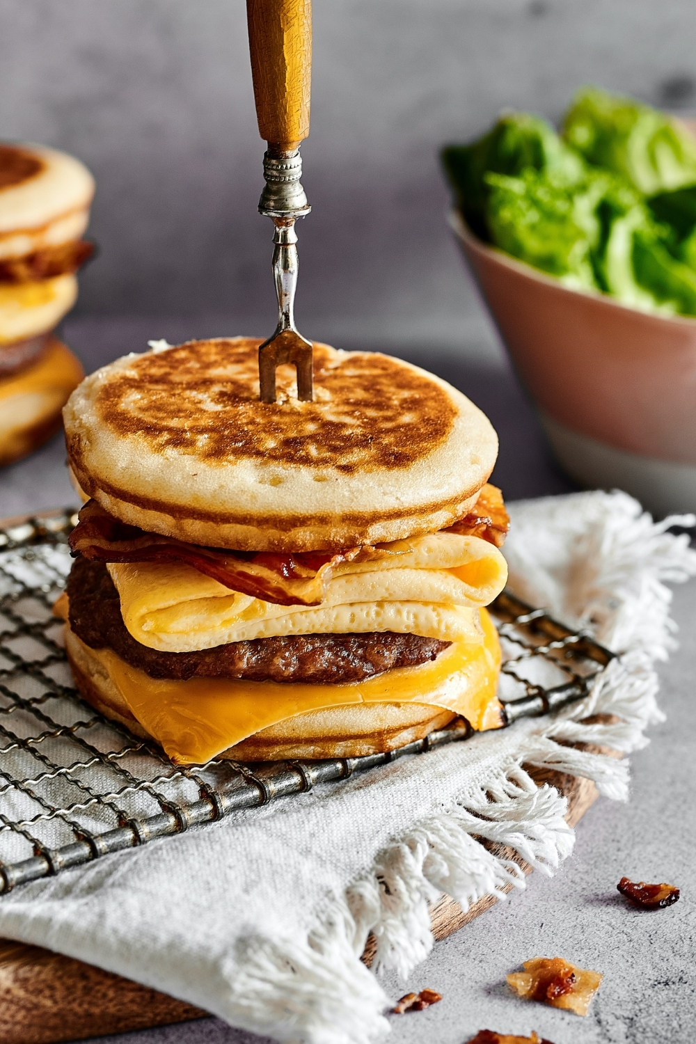 A McGriddle with a pitch fork through sticking out of the top of it. The McGriddle is on a wire rack that is on a white table cloth covering a wooden board on a gray counter. Behind it is part of another McGriddle and to the right of that is part of a bowl of lettuce.