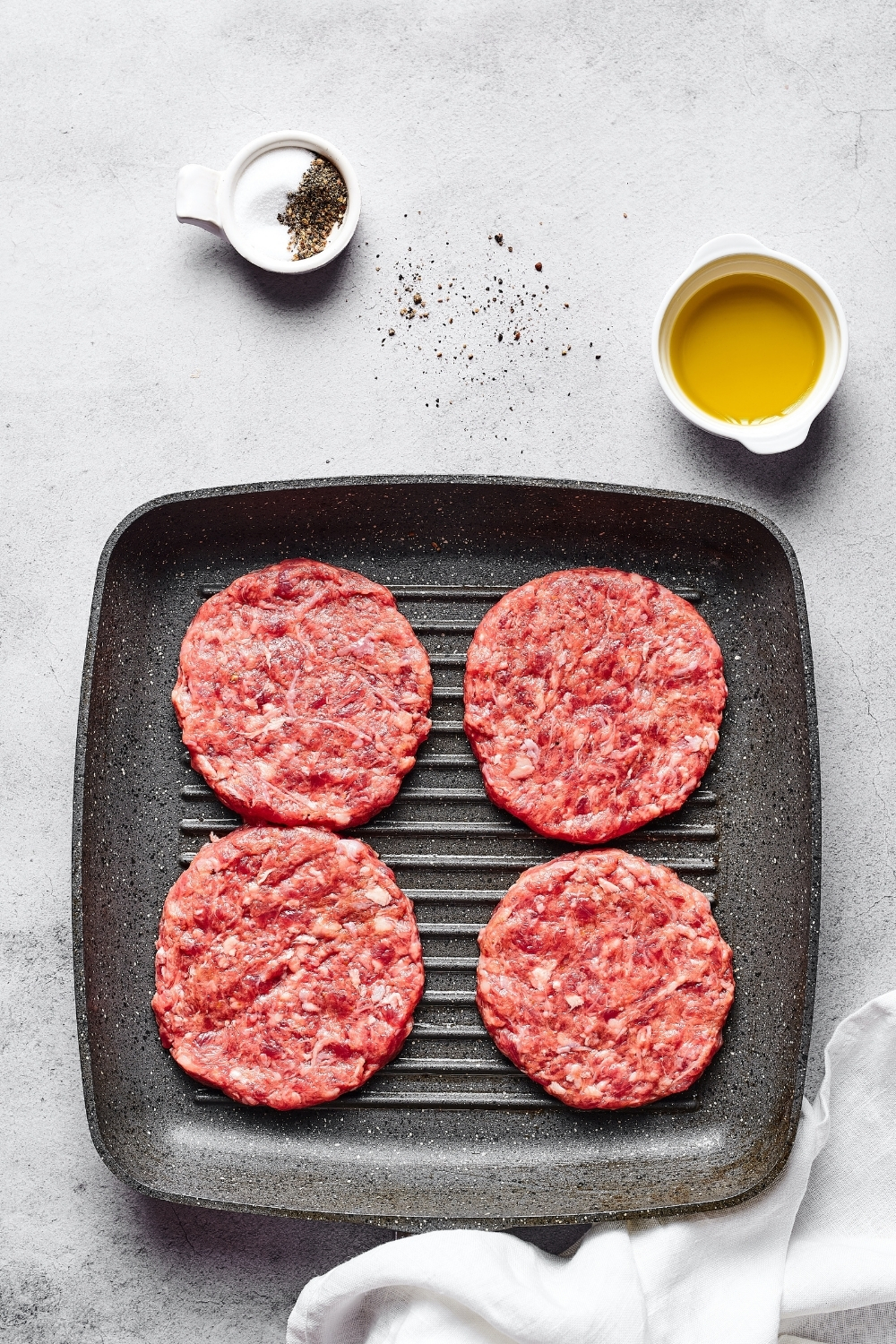 A square grill pan with four meat patties on it. The pan is on a white counter in behind it is a small white bowl of olive oil and a smaller white bowl of salt and pepper.