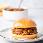 A sloppy Joe on a piece of parchment paper on a gray plate. The gray plate is on a white counter behind it is a white bowl filled with sloppy Joes.