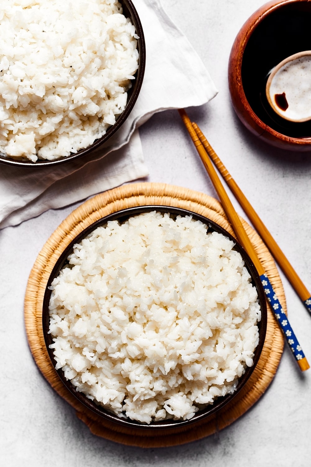 An overhead view of sushi rice and a black bowl and a circular wooden placement. To the right of it is a pair of chopsticks and in front of that part of a wooden bowl fill the soy sauce. Next to the wooden bowl as part of a black bowl filled with sushi rice on a white tablecloth.