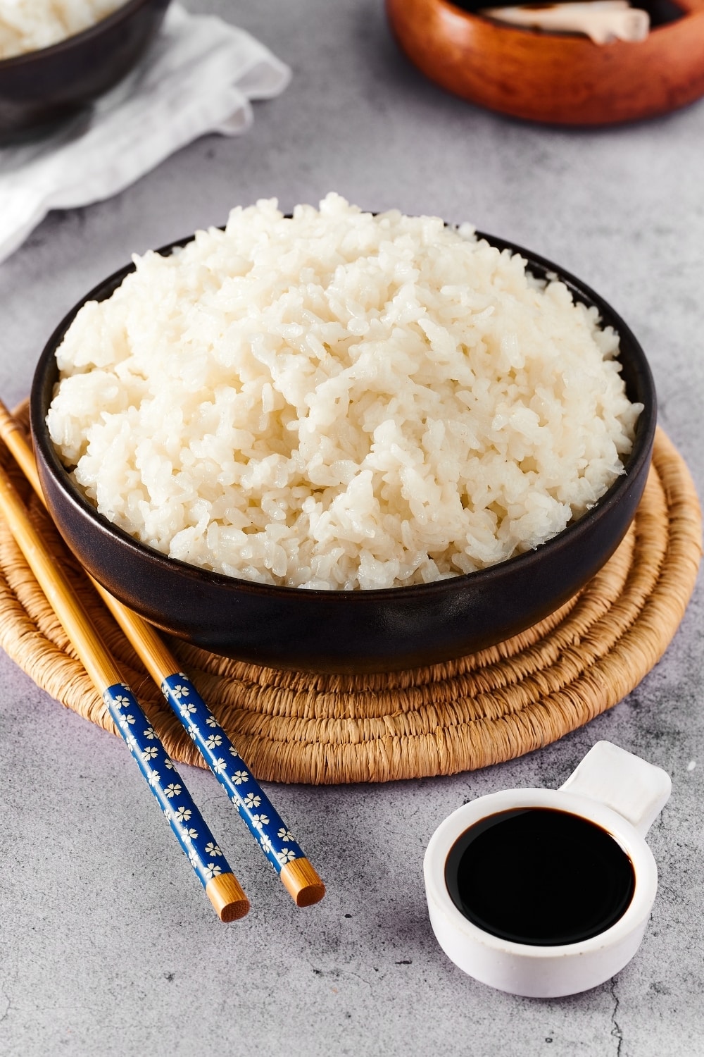 Instant pot sushi rice and a black bowl and a straw circular placemat. To the left of the bowl on the placemat is a pair of chopsticks and in front of the placemat as a small white bowl filled with soy sauce.