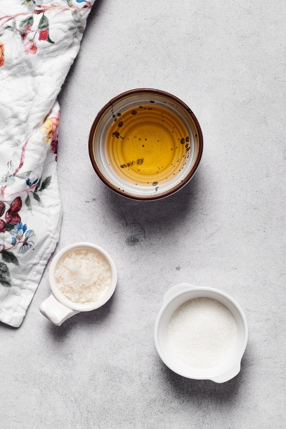 Hey small bowl of rice vinegar, a small cup of sugar, and a small bottle of sea salt on a white counter.