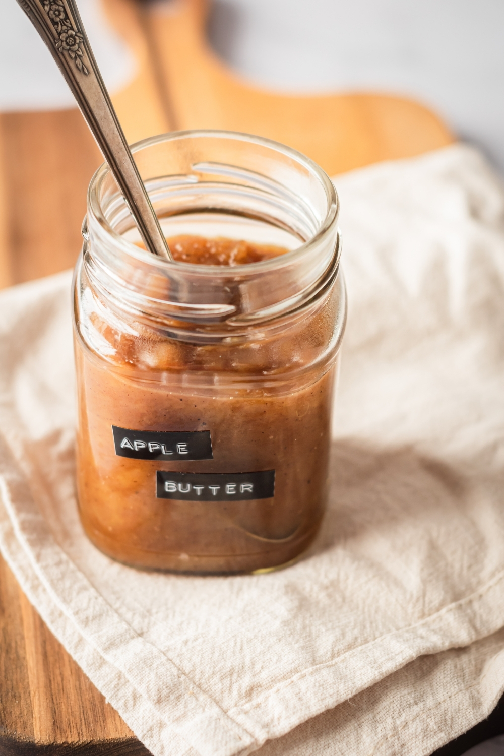 Wooden board with a tablecloth on top with a glass jar of apple butter on it. The glass jar has apple butter labeled on it and there's a spoon submerged in it.