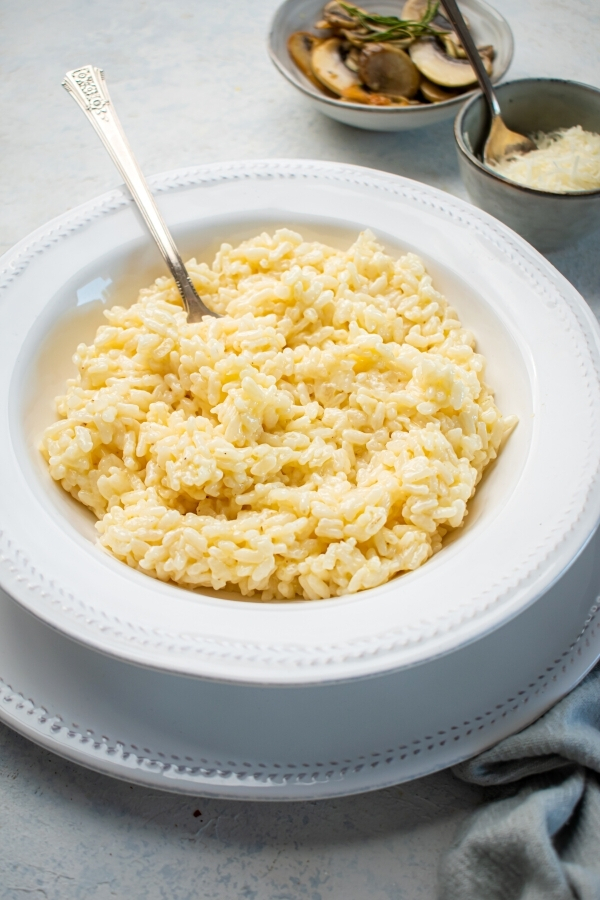 Gray counter with a white plate on it and a white bowl on top filled with risotto. The spoon is submerged in the risotto and behind the bowl is a small bowl of Parmesan cheese and a small bowl of mushrooms.