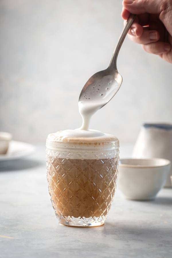 A gray counter with a cup of cold brew with vanilla sweet cream cold foam on top. The hand is holding a spoon over the glass with cold foam dripping from the spoon onto the top of the glass.
