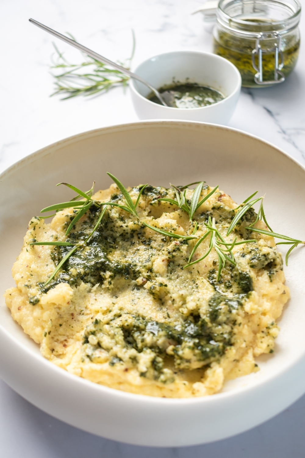Cheesy polenta in a White bowl on the white counter. Behind it is a small white bowl filled with pesto and a glass jar of pesto right behind that.