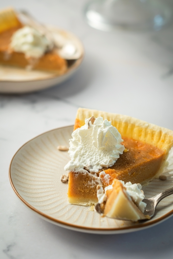 A slice of pumpkin custard pie on a white plate. A spoon has a piece of the front of the pumpkin pie in it.