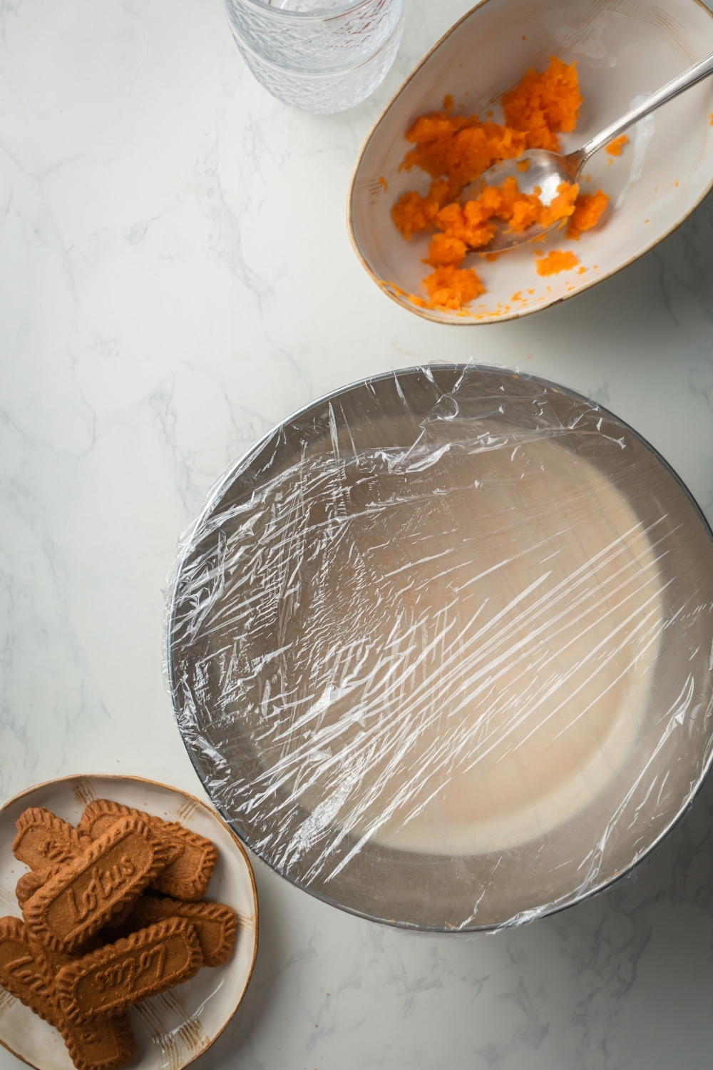 A white counter with a plate of ginger snaps, a silver bowl with pumpkin mousse covered in plastic wrap, and a white dish with pumpkin purée and a spoon in it.