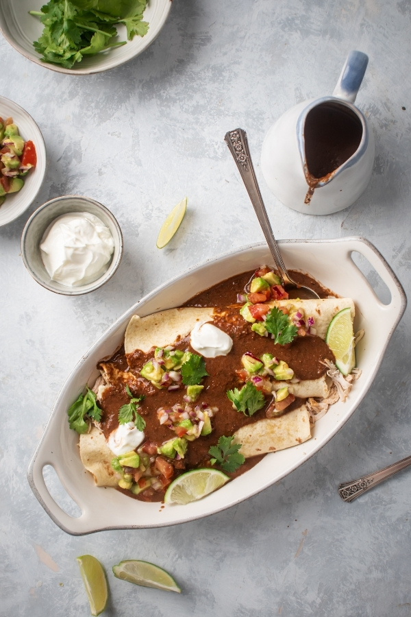 White oval dish on top of a gray counter that is filled with chicken enchiladas. There's a spoon submerged in the enchiladas behind the dishes a small white cup of sour cream and a picture of enchilada sauce.