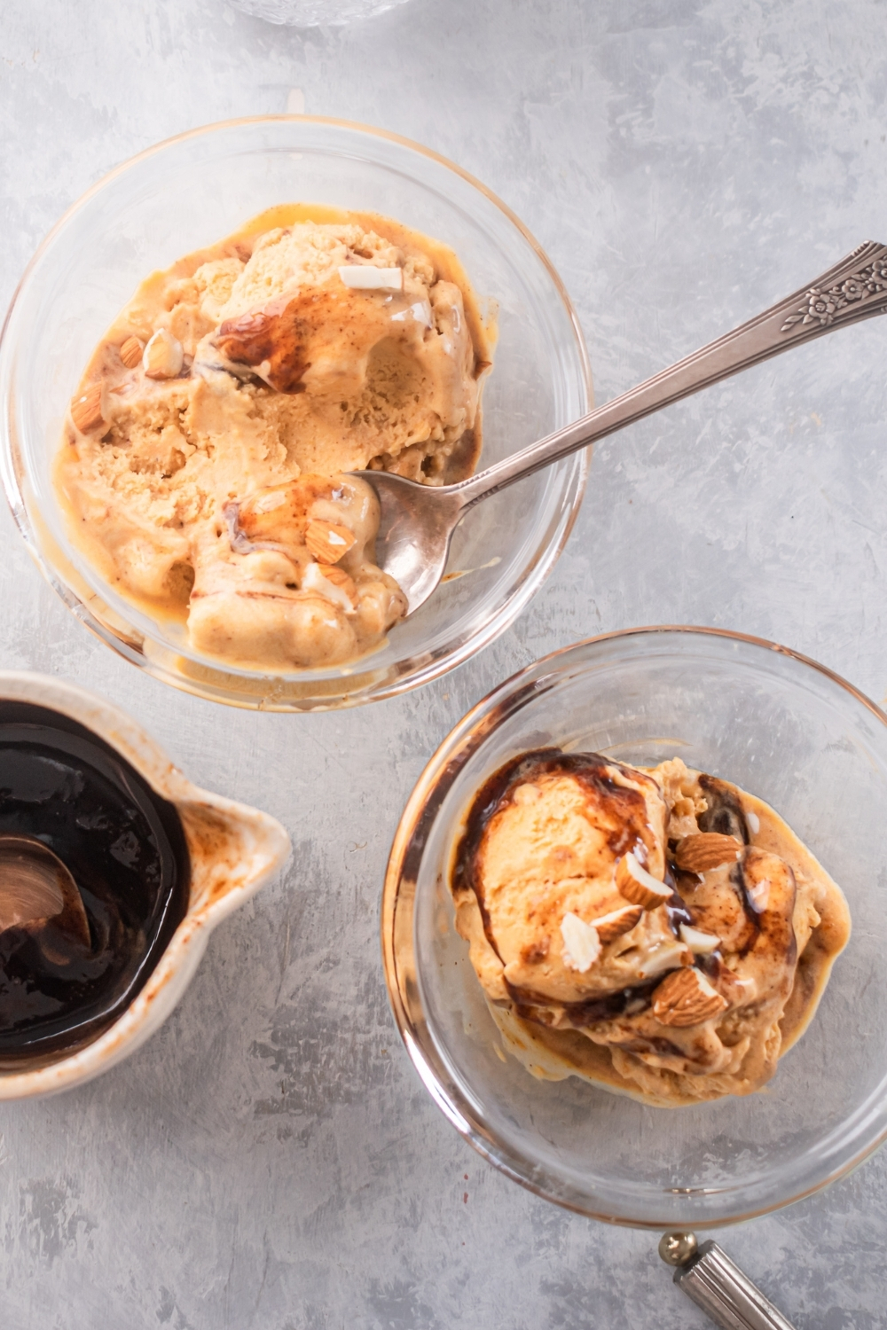 Two glass bowls filled with pumpkin ice cream on a gray counter. The bowl in the back has a spoon submerged in the pumpkin ice cream and to the left of the two bowls as part of a glass pitcher with cinnamon syrup in it.