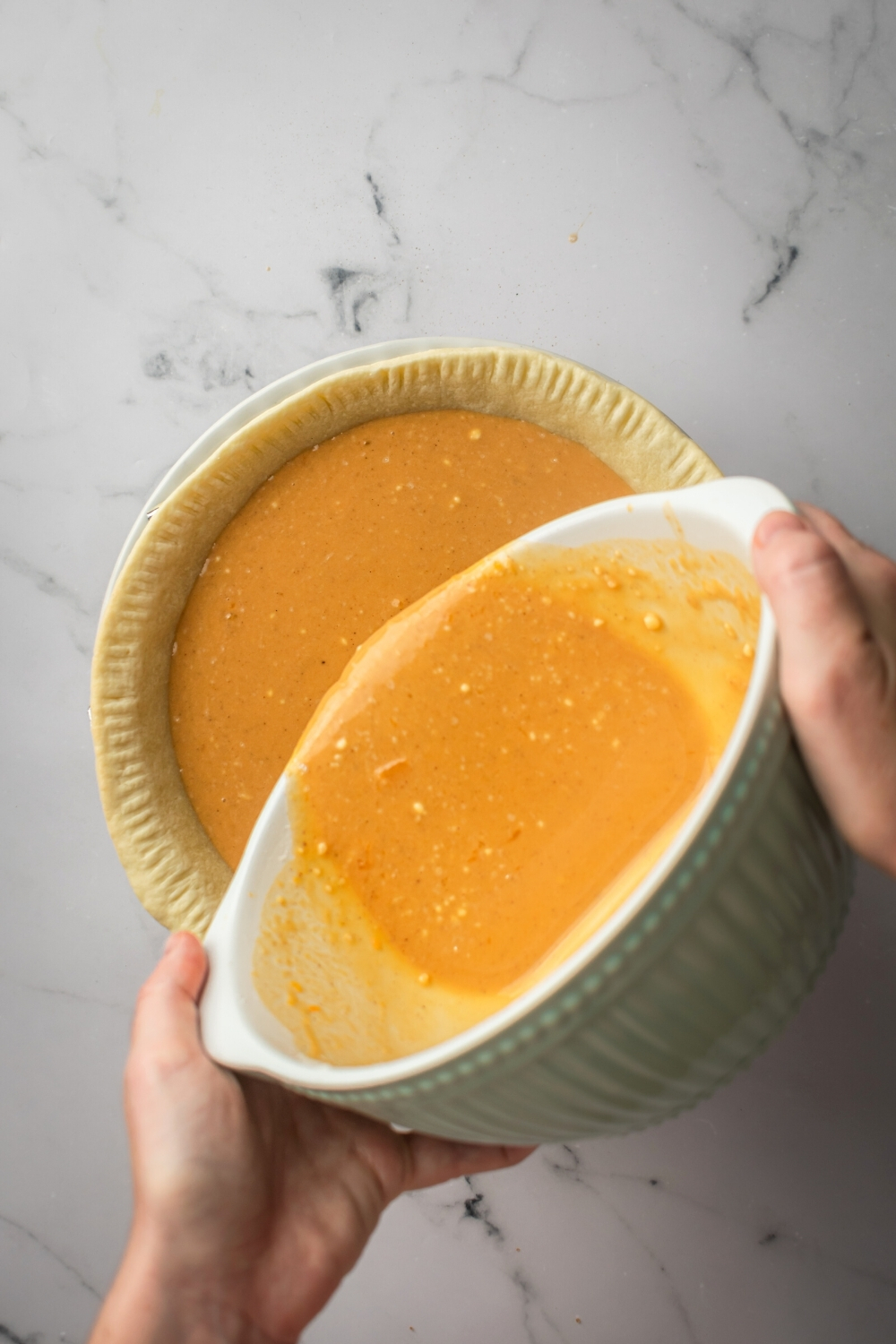 Two hands holding a white bowl pouring pumpkin custard pie filling into a pie crust.