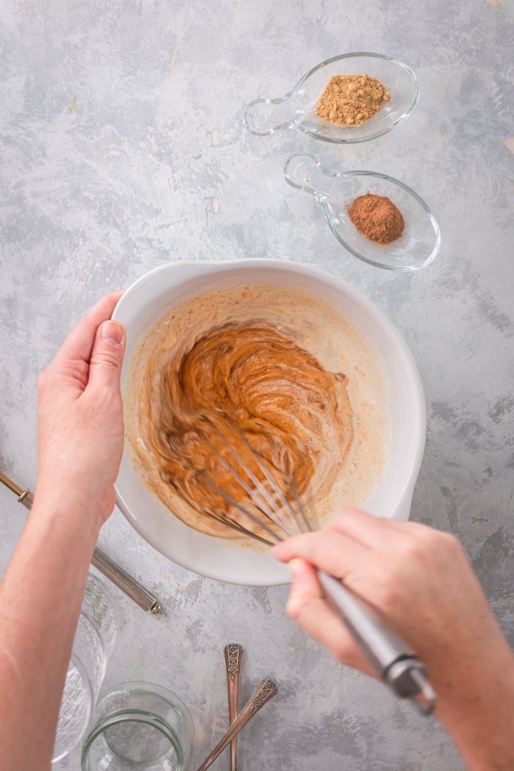 A white bowl on a gray counter with pumpkin ice cream ingredients in it. There is a hand holding a whisk mixing the ingredients together.