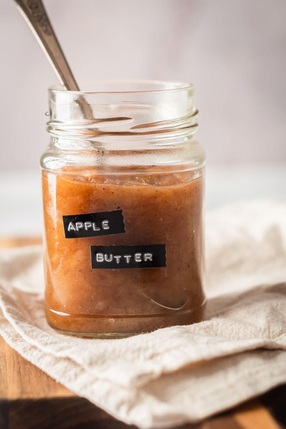A glass jar that is filled with apple butter with a spoon in it on top of a white table cloth. There's a spoon submerged in the jar of apple butter.
