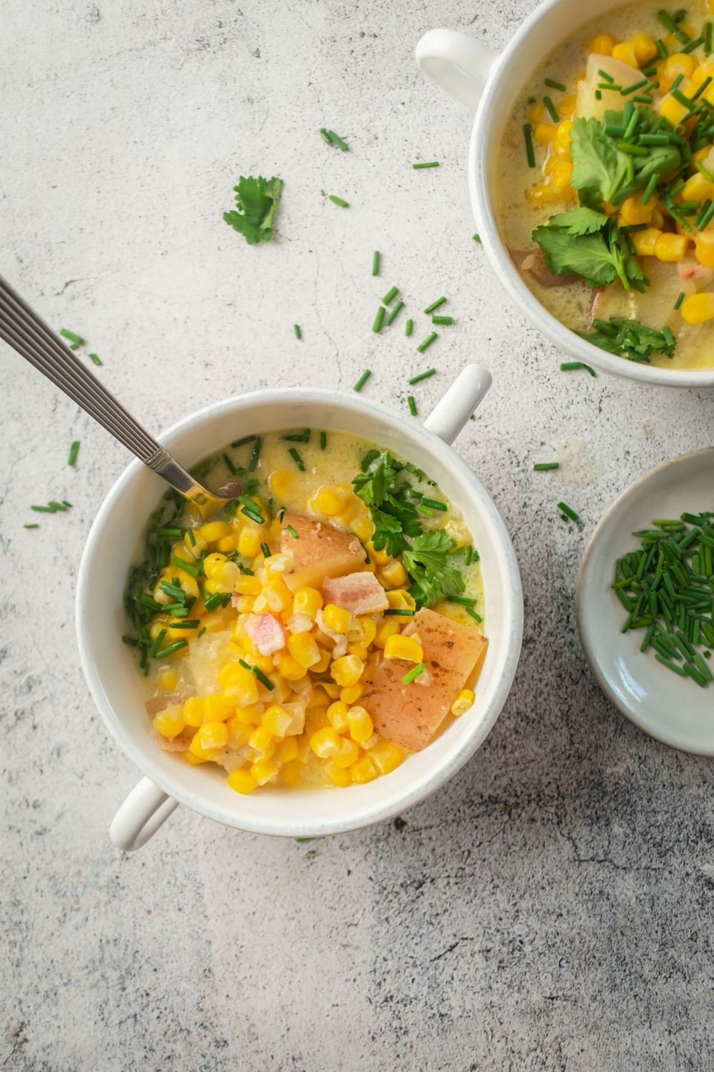 A white counter with a white bowl on top of it filled with corn chowder with a spoon in it. To the back right of it is part of another wipe all filled with corn chowder and in front of that part of a white dish with chives on it.