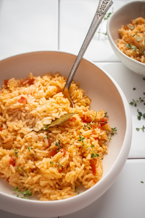 Spanish rice with a spoon in it all in a white bowl. The ball is on white tile and behind it is a small part of another white bowl filled with Spanish rice.