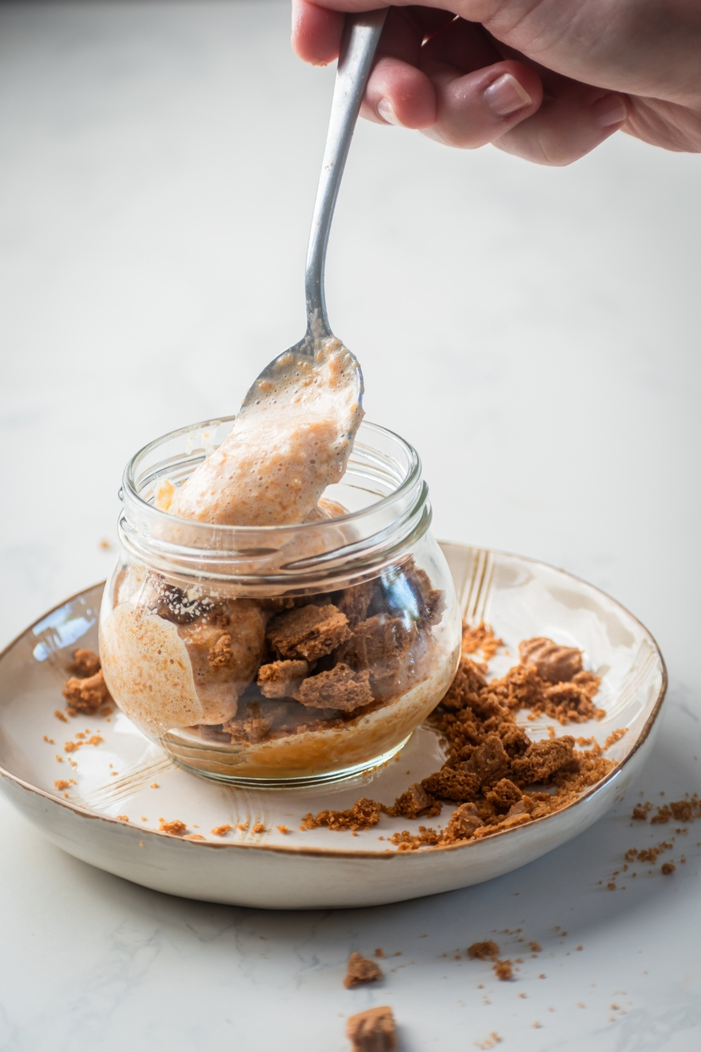 A white plate on a white counter with a glass jar filled with broken ginger snaps on top of it. There is a hand spooning pumpkin mousse into the jar.