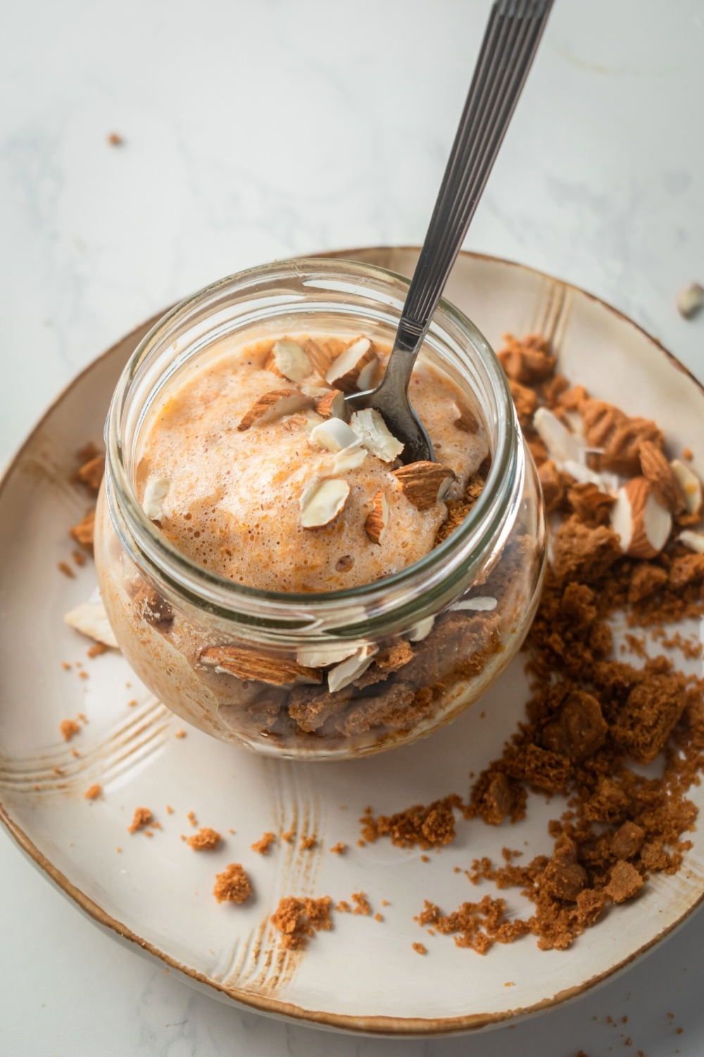 A glass jar filled with pumpkin mousse, almonds, and ginger snaps with a spoon submerged in it. Glass jar is on a white plate with gingersnap crumbles on it.