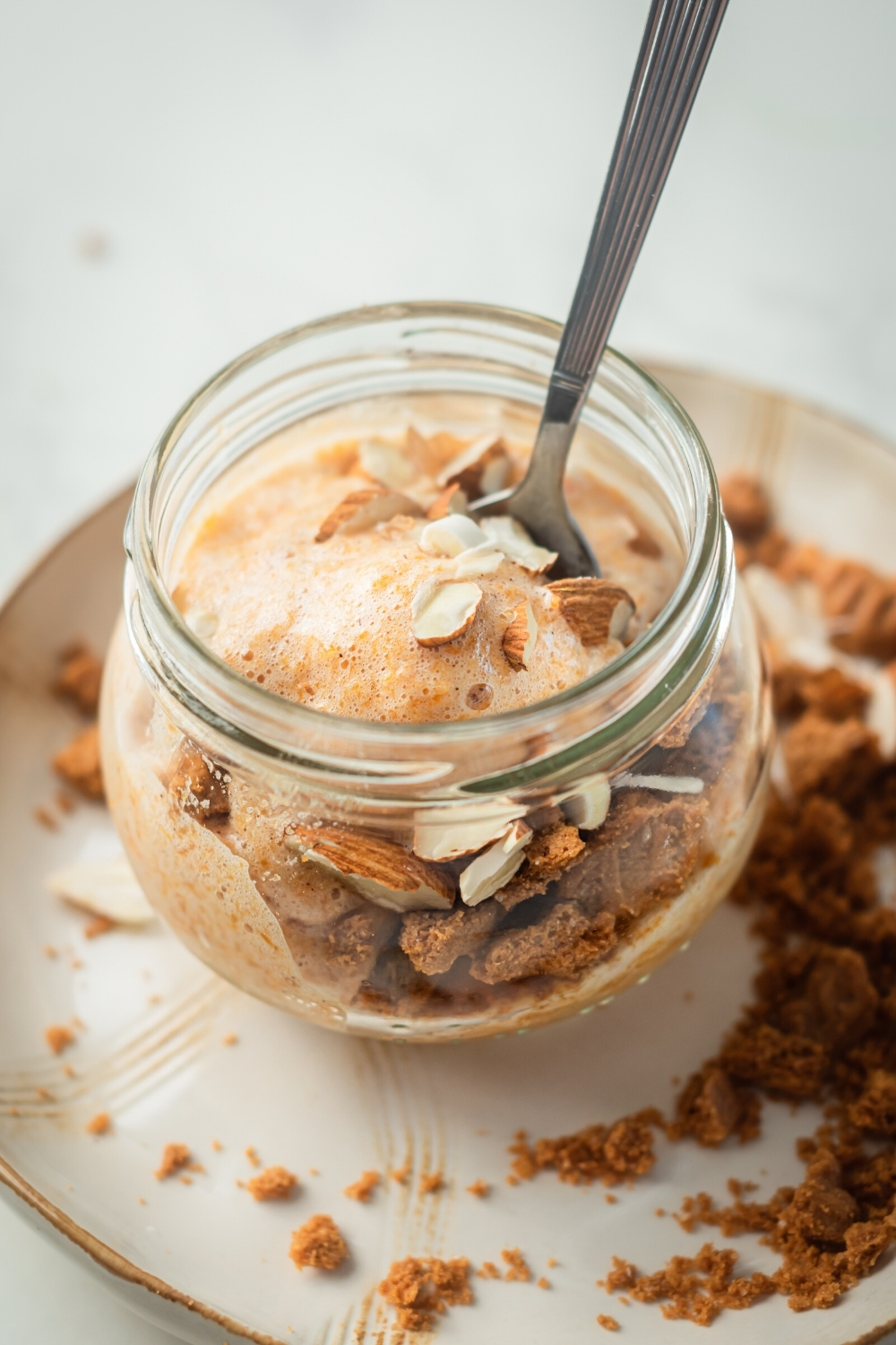 A glass jar filled with pumpkin mousse, almonds, and ginger snaps. The jar of moose is on a white plate and it is surrounded by ginger snap crumbles on the plate.
