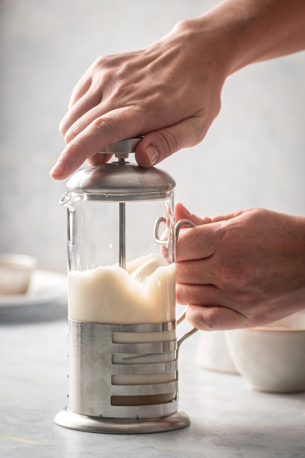 A French press filled with sweet cream cold foam on a gray counter. One hand is holding the handle of the French press and the other is holding the top of it.