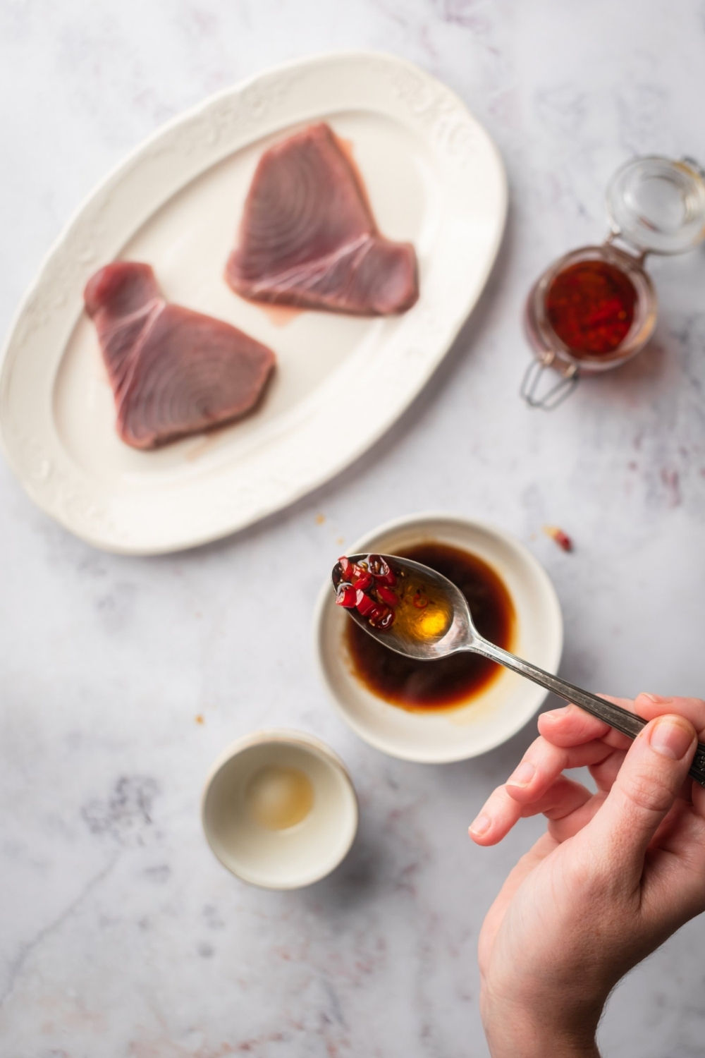 A hand holding a spoon with chili peppers on it over a grey counter with a plate of two tuna steaks, a glass jar of chili peppers, and a bowl of soy sauce on it.
