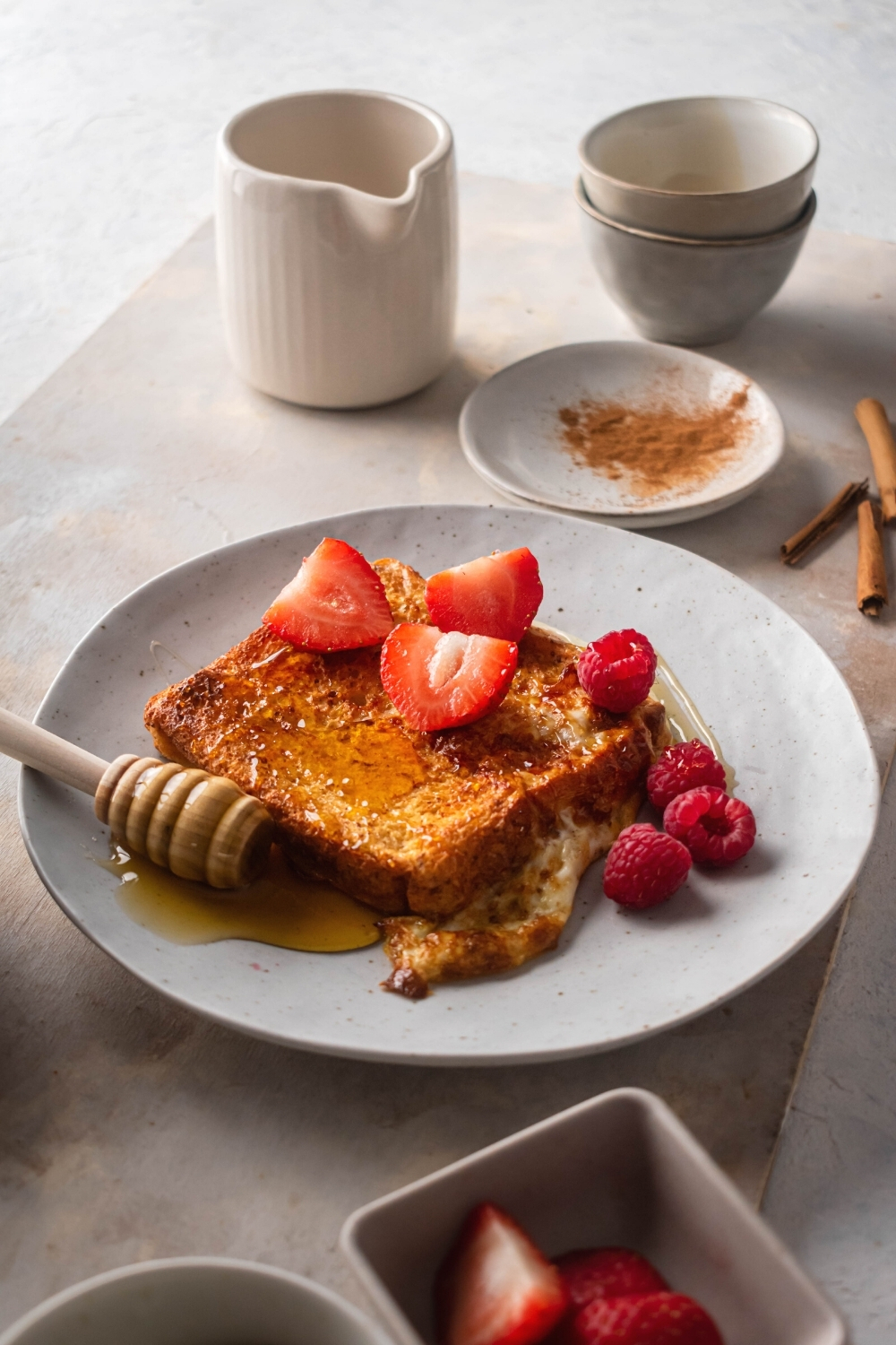 A white plate with a piece of French toast on it. There are some raspberries next to the French toast and some sliced strawberries on top. Behind it is a small white plate with cinnamon on it, a white pitcher, and two small white bowls stacked on top of each other.