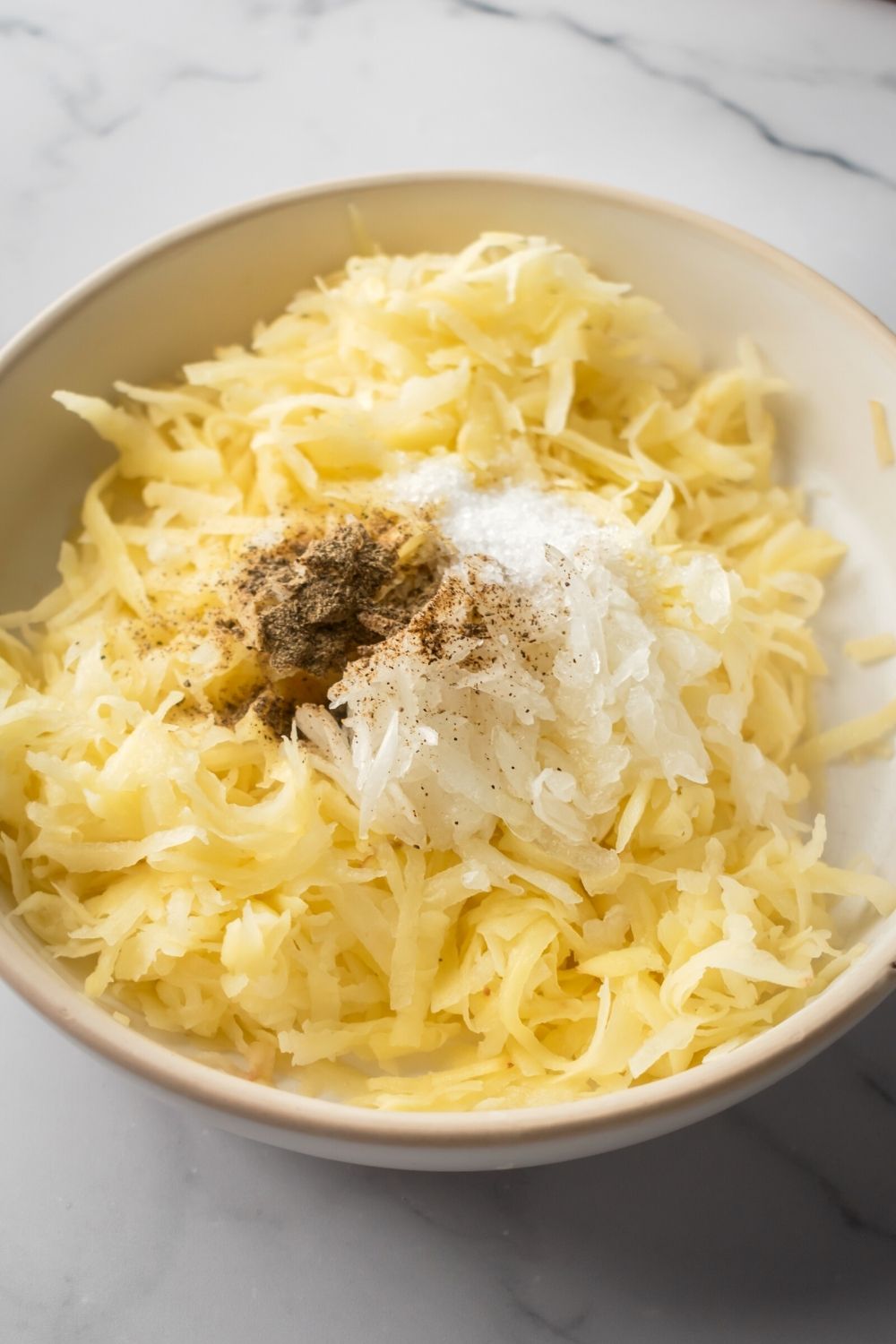 White bowl with shredded potatoes, onions, and salt and pepper in it.