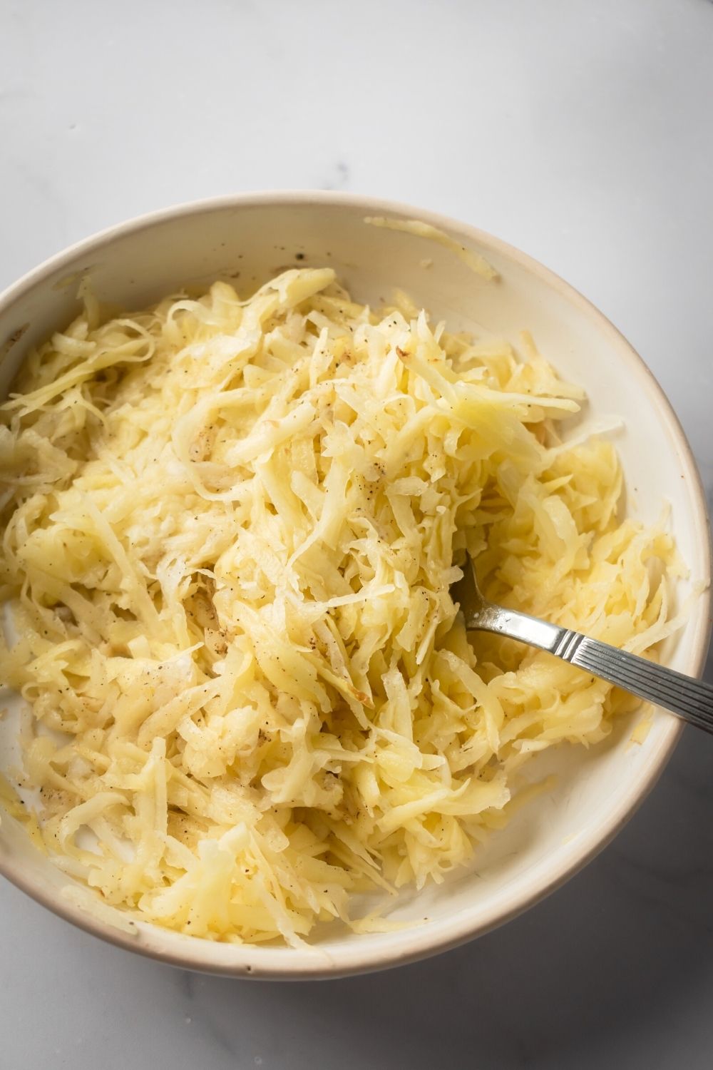 A white bowl filled with shredded potatoes with a fork submerged in the potatoes.