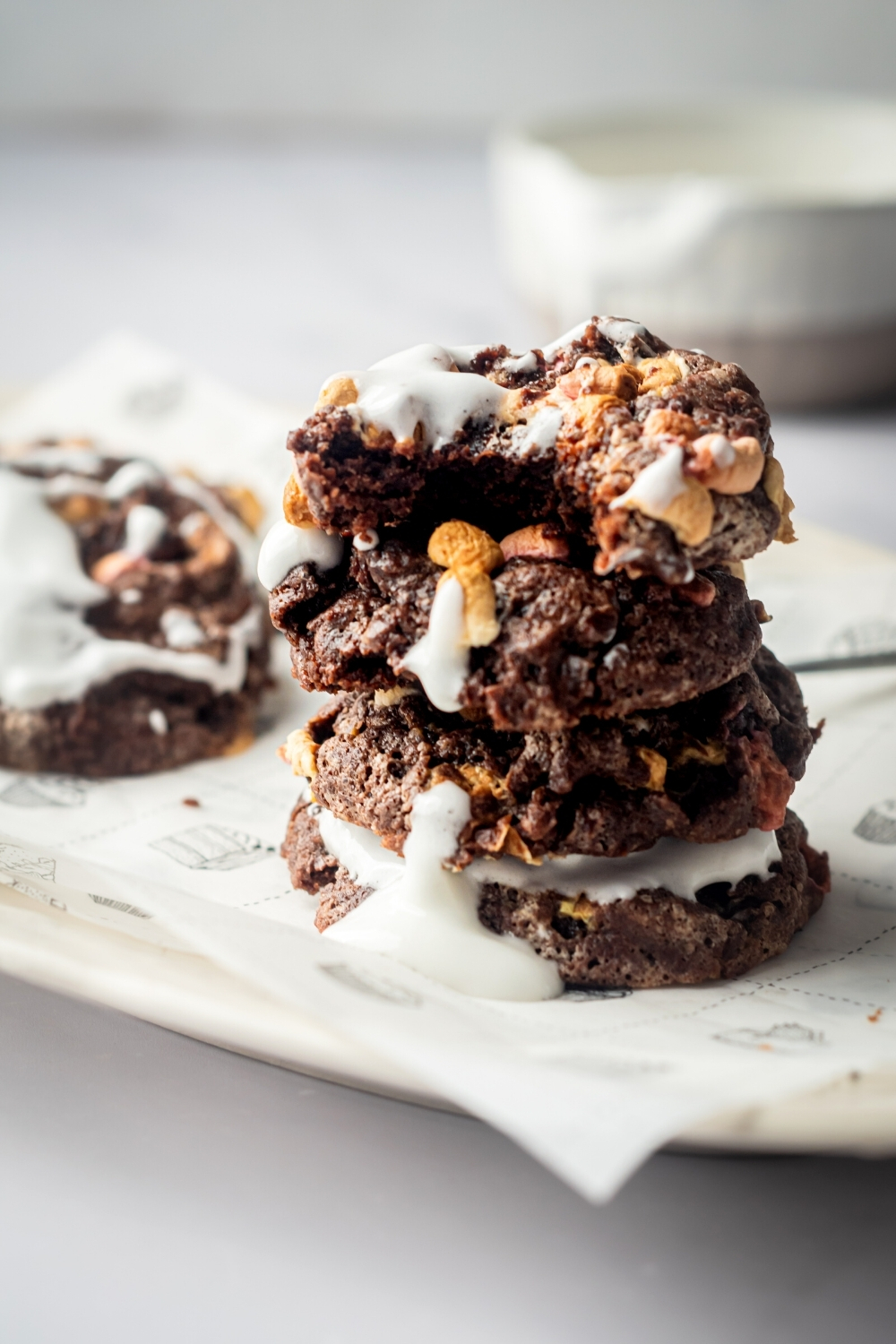 A stack of four chocolate marshmallow cookies on a piece of parchment paper on a white plate. The top marshmallow cookie has a bite out of the front of it.