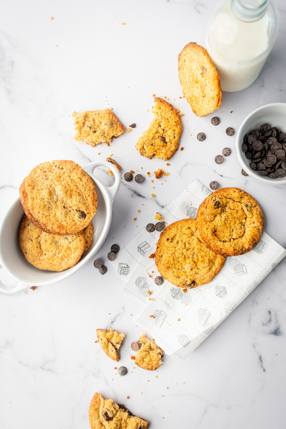 White counter with two chocolate chip cookies on a piece of parchment paper, a white bowl with chocolate chip cookies in it, a few pieces of chocolate chip cookie, and chocolate chips on it.