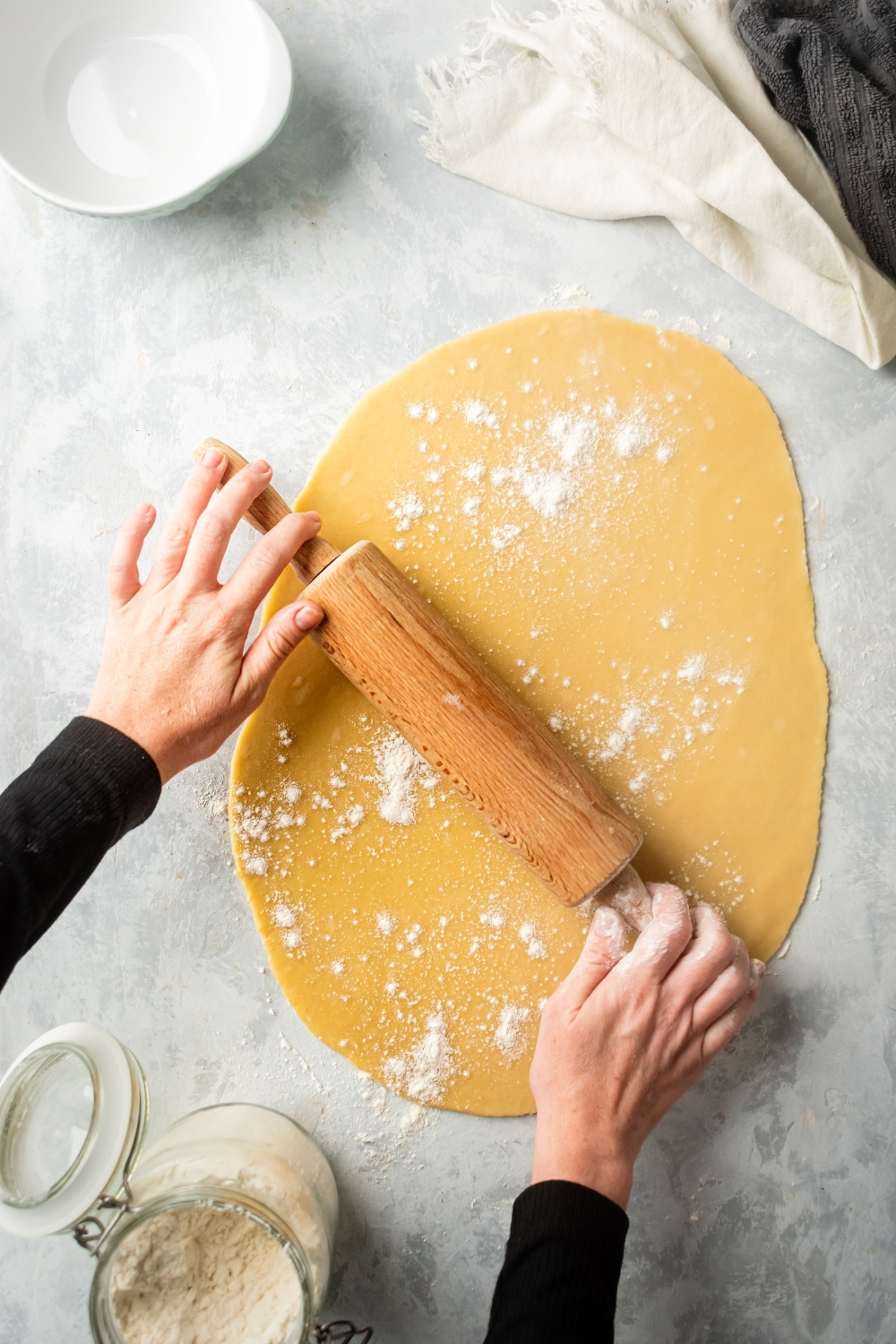 A thin piece of rolled out and empanada dough. Two hands are holding a roller on top of the dough and there is a little flour on the dough.